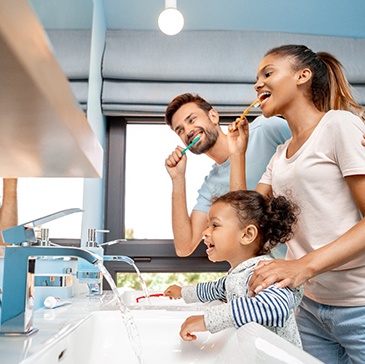 A man, woman, and little girl brushing their teeth together at bathroom sinks
